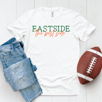White t-shirt with 'Eastside' in green block letters, 'the best side' in orange script, blue jeans, and football on a white wooden surface